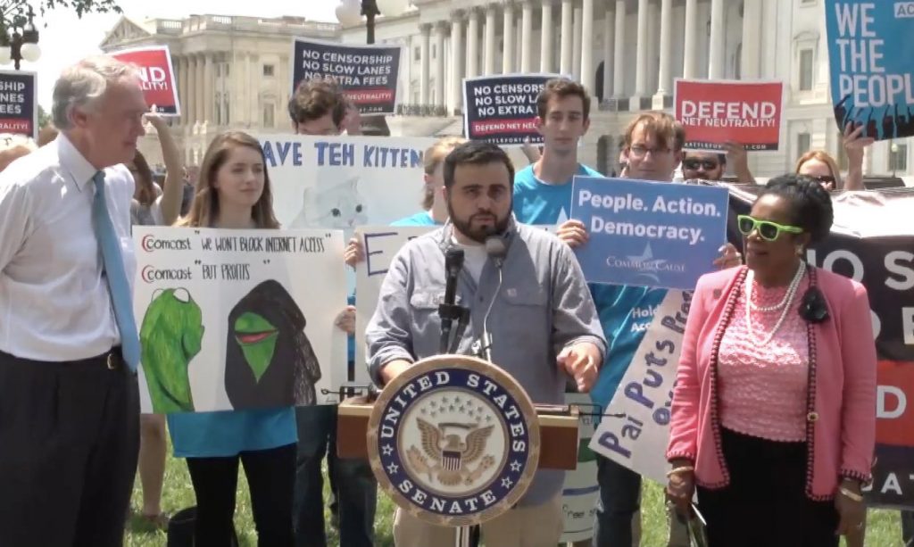 Steven Renderos speaks on net neutrality in front of the US Senate. Protestors hold "Save net neutrality" signs in the background.