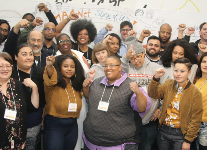 A group photo of the MediaJustice Network in 2018 in Oakland, CA. Many smiling faces with hands raised in a show of collective power.