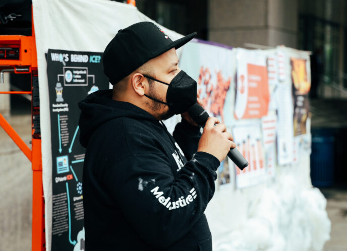MediaJustice Field Organizer Danny Cendejas, wearing a black cap, mask, and MediaJustice hoodie speaks into a mic in front of a public art installation telling the story of Amazon & AWS's global impact of state violence