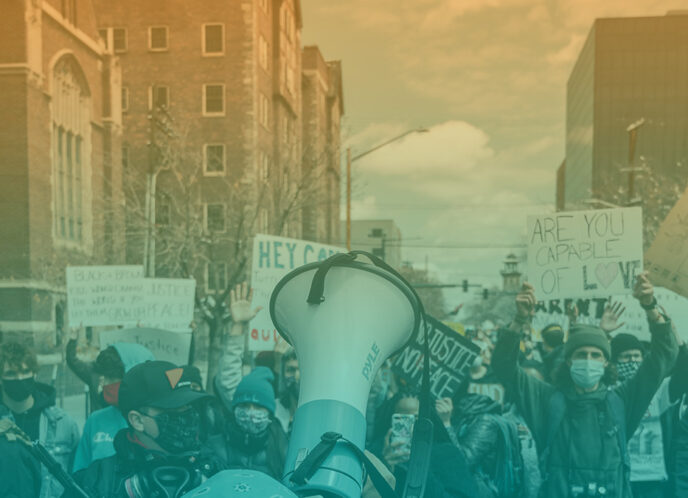 color-washed photo depicts a protest where masked people hold signs, a megaphone is raised in the foreground, an armored person patrols the edge and buildings surround the frame