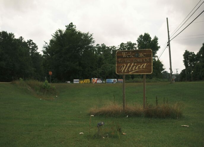 PHOTO: a field and treeline by a rural Mississippi road, with a sign reading "Welcome to the town of Utica" in the foreground