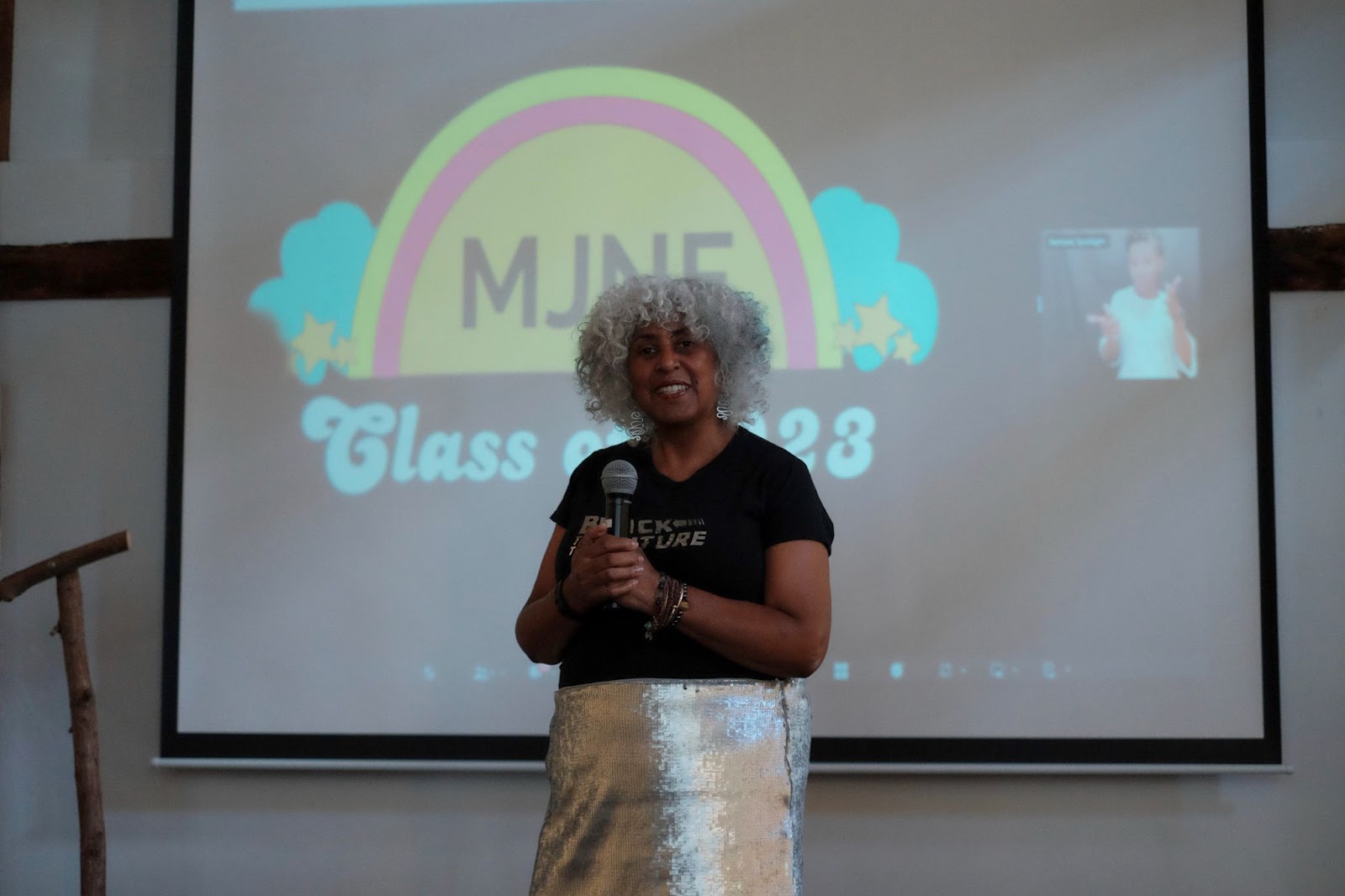 Keynote speaker and movement communications trailblazer Makani Themba speaks in front of a projector screen reading 'MJNF Class of 2023', wearing a shirt that reads 'Black to the Future'